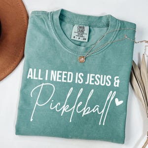 Puede incluir: Una camiseta verde azulada con el texto "All I need is Jesus & Pickleball" en letras blancas con un peque&ntilde;o coraz&oacute;n blanco.