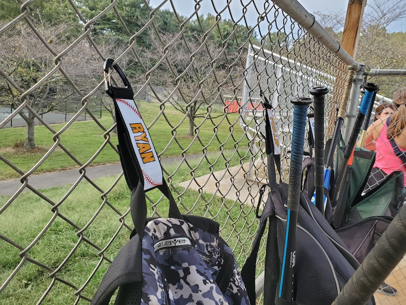 May include: A black backpack with a camouflage pattern and a black and white baseball strap with the name "RYAN" printed in orange letters. The strap is attached to a chain link fence.