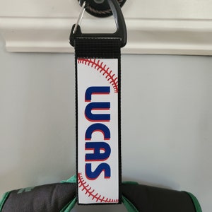 May include: A black and white personalized luggage tag with a baseball graphic and the name "LUCAS" printed in blue letters.