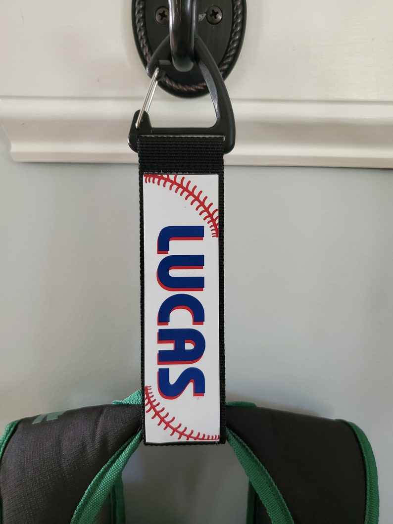 May include: A black and white personalized luggage tag with a baseball graphic and the name "LUCAS" printed in blue letters.