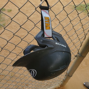 May include: A black baseball helmet with a white logo hanging on a chain link fence. A white and yellow lanyard with the name "RYAN" is attached to the helmet.