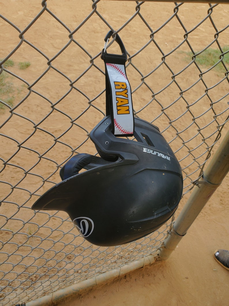 May include: A black baseball helmet with a white logo hanging on a chain link fence. A white and yellow lanyard with the name "RYAN" is attached to the helmet.