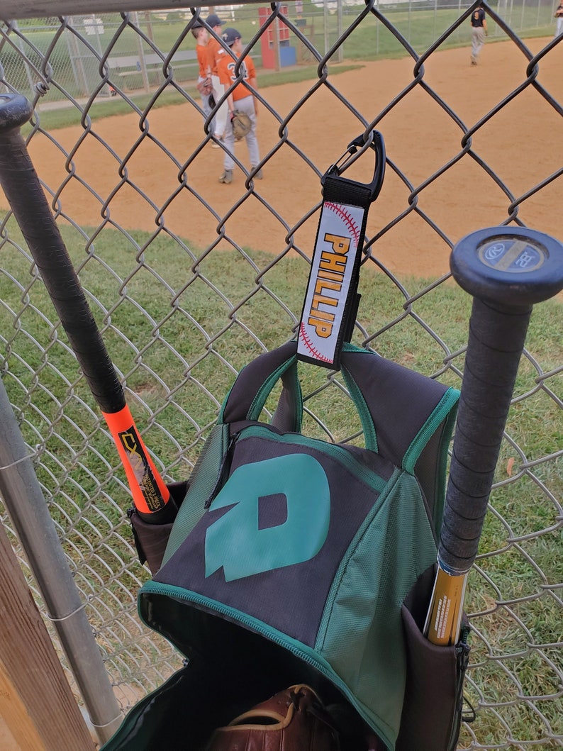 May include: A green and black baseball backpack with a large letter "D" on the front. Two baseball bats are in the pockets of the backpack. A black and white baseball keychain with the name "Phillip" is hanging from the backpack.