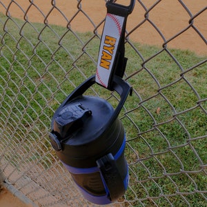 May include: A blue and black water bottle with a black carabiner clip attached to a white strap with the name "RYAN" printed in orange letters.