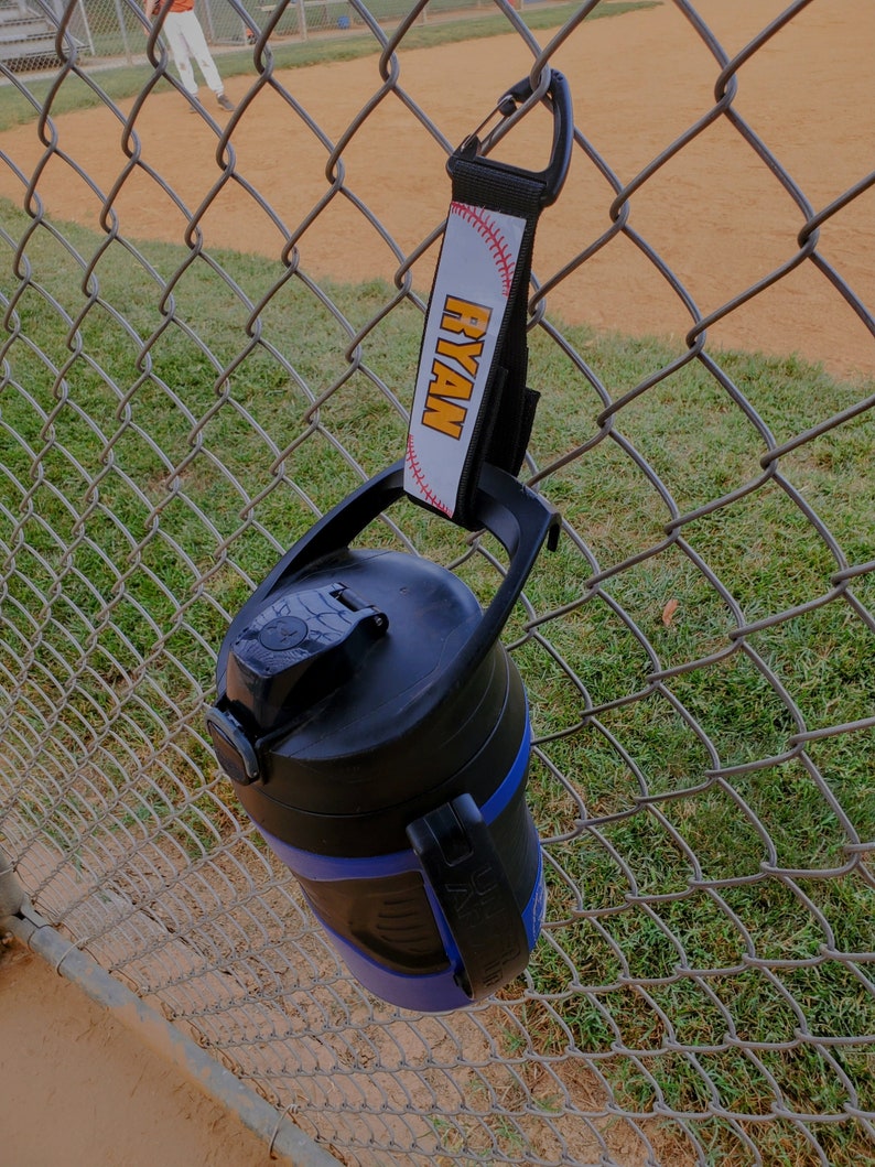 May include: A blue and black water bottle with a black carabiner clip attached to a white strap with the name "RYAN" printed in orange letters.