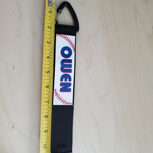 May include: Black and white baseball-themed keychain with a black strap and a silver clip. The keychain has the name "Owen" printed in blue letters on a white baseball.