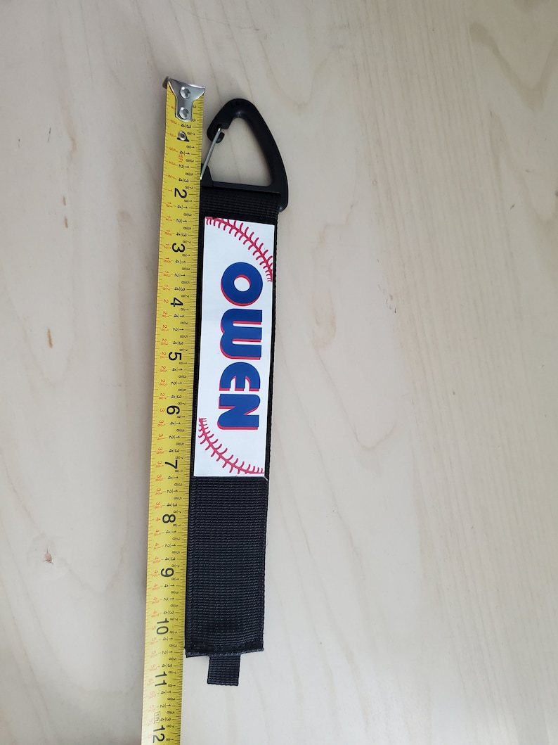 May include: Black and white baseball-themed keychain with a black strap and a silver clip. The keychain has the name "Owen" printed in blue letters on a white baseball.