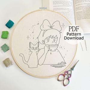 May include: Embroidery hoop featuring a black and white line drawing of a girl drinking from a teacup, with a cat beside her. The text "PDF Pattern Download" is visible. Embroidery floss in shades of green and teal, along with scissors and needles, are also present.