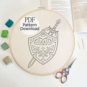 May include: An embroidery hoop with a beige fabric featuring a black line drawing of a sword and shield design. The text "PDF Pattern Download" is in a white circle. Colourful embroidery floss and scissors are also in the image.