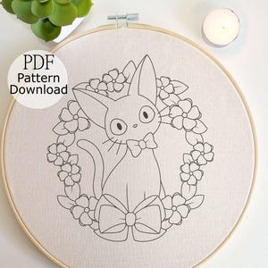 May include: Embroidery hoop featuring a black and white cat design surrounded by flowers and a bow. The text "PDF Pattern Download" is visible. The design is on a beige fabric, set in a wooden hoop.