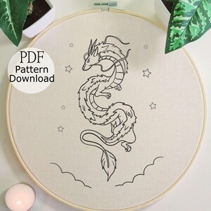 Może przedstawiać: Tamborek z czarnym rysunkiem smoka, gwiazd i chmur. Tekst "PDF Pattern Download" znajduje się w białym okręgu. Tamborek otoczony jest roślinami i zapaloną świecą.
