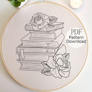 May include: An embroidery hoop containing a cream-colored fabric with a black line art design of a stack of four books adorned with two roses. The text PDF Pattern Download is printed in black on the right side of the fabric within the hoop.