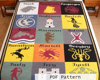 Puede incluir: Una colorida manta con los emblemas de las casas de una serie de fantasía. El diseño incluye cuadrados con nombres de casas y emblemas como un lobo, un león y un dragón. La manta tiene un borde negro y dorado.