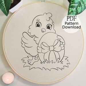 May include: An embroidery hoop with a black and white line drawing of a cartoon chick and an Easter egg with a bow. The text "PDF Pattern Download" is in a white circle. The design is on a cream-colored fabric.