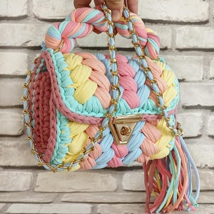May include: A pastel-colored handbag with a braided design. The bag features a gold chain strap and clasp, with a tassel of matching colors. The handle is also braided, and the bag is made of soft, colorful fabric.