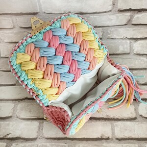 May include: A handmade, open handbag with a woven design in pastel colors. The bag features a woven pattern of pink, blue, and yellow fabric strips. The edges are trimmed with a braided border and a tassel of matching colors. A gold-colored clasp is visible.