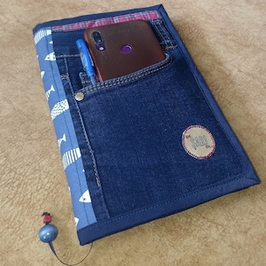 Peut inclure: Un carnet en jean bleu avec un motif de poisson blanc et un accent en tissu à carreaux rouge et blanc. Un téléphone portable et un stylo sont rangés dans une poche sur la couverture avant. Le carnet a une bordure en cuir brun et une petite étiquette brune avec le texte "DE. SARU. OL."