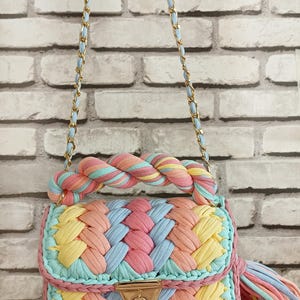 May include: A colorful, handmade handbag with a braided design. The bag features a pastel color palette of pink, yellow, blue, and peach. It has a gold chain strap and a matching tassel. The bag is made of a soft, woven material.