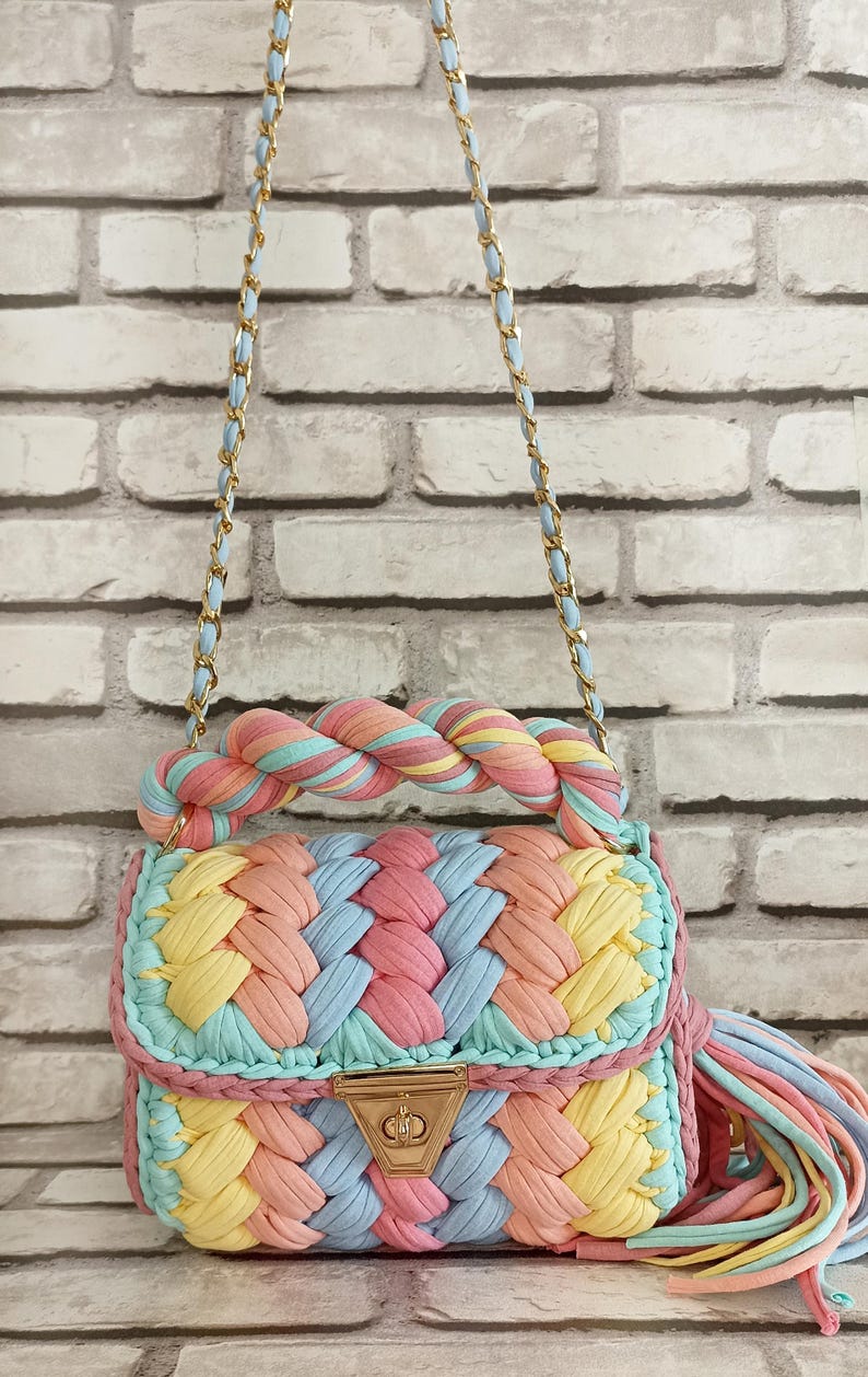May include: A colorful, handmade handbag with a braided design. The bag features a pastel color palette of pink, yellow, blue, and peach. It has a gold chain strap and a matching tassel. The bag is made of a soft, woven material.
