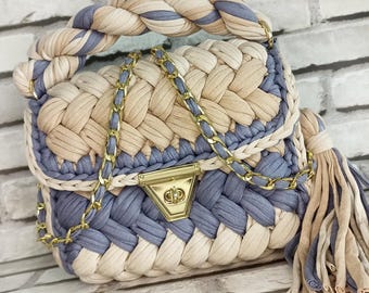 May include: A woven handbag with a beige and blue color scheme. The bag has a braided handle and a gold chain strap. The bag has a gold clasp and a tassel.