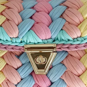 May include: Close-up of a pastel-colored handbag with a woven design. The bag features a gold-colored clasp and is made of braided fabric in shades of pink, blue, yellow, and green. The overall aesthetic is soft and summery.