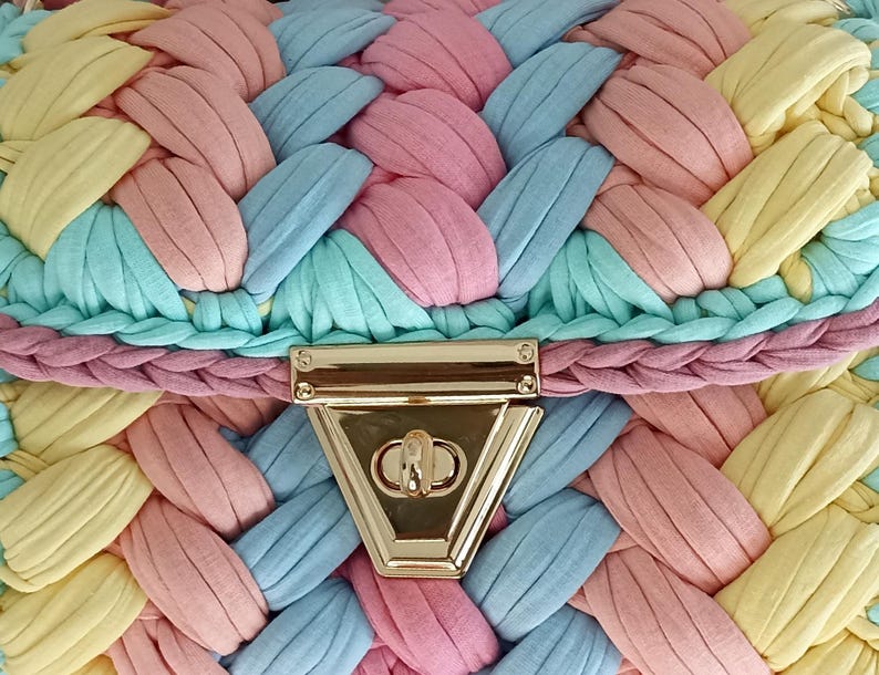 May include: Close-up of a pastel-colored handbag with a woven design. The bag features a gold-colored clasp and is made of braided fabric in shades of pink, blue, yellow, and green. The overall aesthetic is soft and summery.