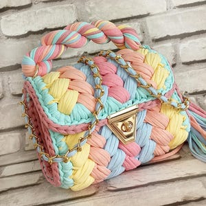 May include: A pastel-colored, hand-woven handbag with a braided handle and gold-tone chain strap. The bag features a woven design in shades of pink, yellow, blue, and peach. The clasp is gold-toned. A tassel hangs from the side.