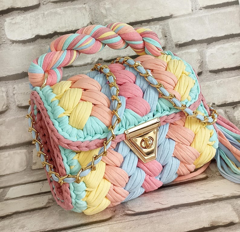 May include: A pastel-colored, hand-woven handbag with a braided handle and gold-tone chain strap. The bag features a woven design in shades of pink, yellow, blue, and peach. The clasp is gold-toned. A tassel hangs from the side.