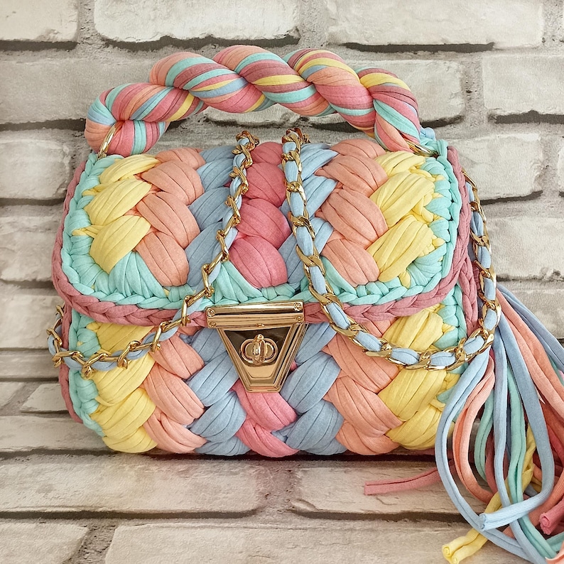 May include: A pastel-colored handbag with a braided design. The bag features a woven pattern in shades of pink, yellow, blue, and peach. It has a twisted handle and gold chain straps. A gold clasp secures the bag, and a tassel hangs from the side.