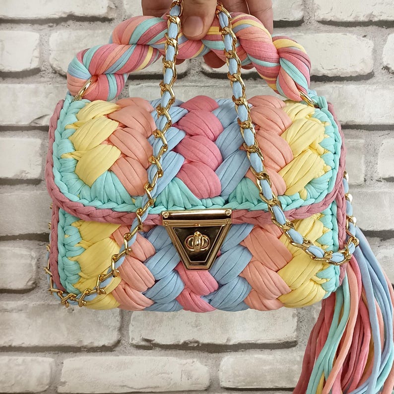 May include: A handmade, colorful handbag with a braided design. The bag features a mix of pastel colors, including pink, yellow, blue, and peach. It has a gold chain strap and a tassel. The bag is held by a hand.