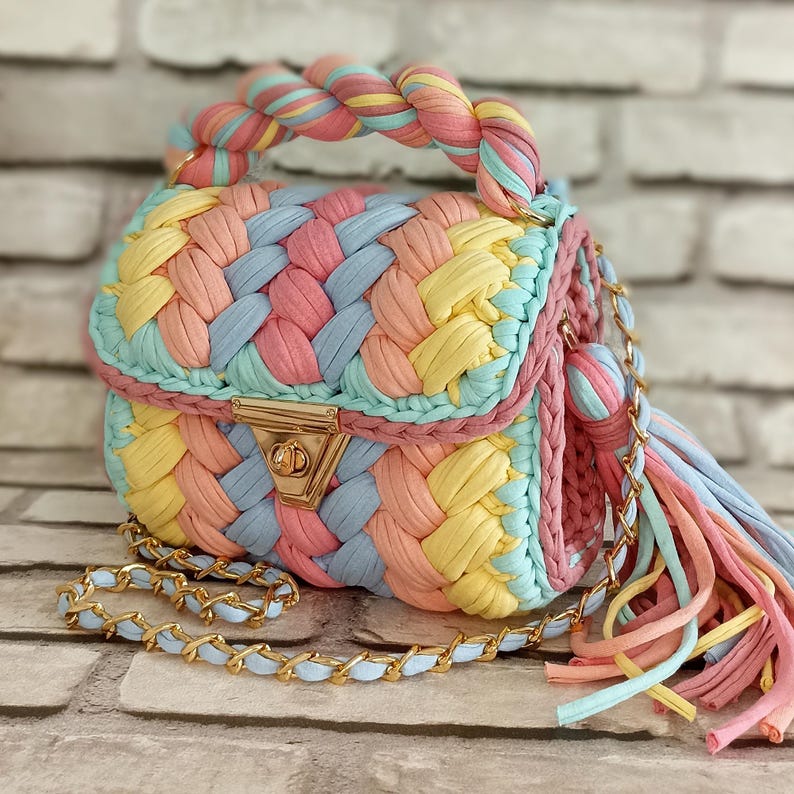 May include: A colorful, woven handbag with a braided handle and a gold-tone clasp. The bag features a pastel color palette of pink, yellow, blue, and green. A gold chain strap and a tassel add to the bag's design.