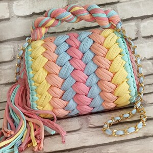 May include: A colorful, woven handbag in pastel shades of pink, yellow, blue, and peach. The bag features a braided handle and a gold chain strap. A tassel of matching colors hangs from the side.