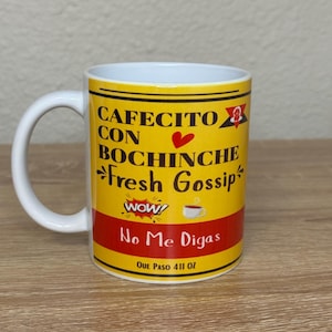 May include: White ceramic coffee mug with a yellow and red design. The mug features the text "CAFECITO CON BOCHINCHE Fresh Gossip" and "No Me Digas". The mug has a white handle and holds approximately 11 oz.