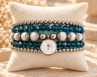 May include: A multi-strand beaded bracelet featuring rows of metallic silver beads, faceted dark teal crystal beads, and smooth white beads. A central white circular charm with a silver cross emblem hangs from the bracelet.