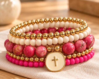 May include: A stack of five beaded bracelets featuring metallic gold spheres, matte white beads, and vibrant pink marbled stone beads. A circular cream-colored charm with a gold cross emblem is attached to the center of the bracelet stack.