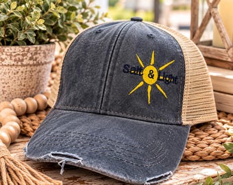 May include: A charcoal gray distressed baseball cap with a tan mesh back. The front panel features an embroidered yellow sun graphic with the text Salt & Light in dark blue lettering. The brim has a frayed edge and visible stitching.