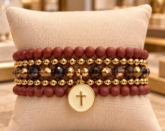 May include: A multi-strand beaded bracelet set featuring matte maroon beads, polished gold-tone metallic beads, and faceted dark brown beads. A circular white enamel charm with a gold-tone cross emblem hangs from the center of the stack.