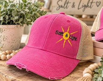 May include: A bright pink baseball cap with a distressed brim and a tan mesh back. The front panel features an embroidered yellow sun graphic with the black text Salt & Light centered across the middle of the sun design.