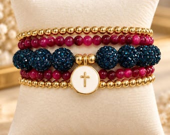 May include: A multi-strand beaded bracelet set featuring gold-tone metallic beads, deep pink smooth beads, and dark blue textured glitter beads. A central white circular charm with a gold-tone cross emblem is attached to the bracelet set.