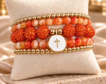 May include: A set of four stacked beaded bracelets featuring gold-tone metallic beads, orange marbled stone beads, and vibrant orange glitter-coated beads. A central white enamel charm with a gold-tone cross emblem hangs from the stack.
