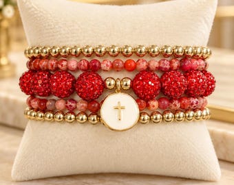 May include: A stack of five beaded bracelets featuring gold-tone metallic beads, red speckled stone beads, and large red crystal-encrusted spheres. A central white circular charm with a gold-tone cross emblem hangs from the bracelet stack.
