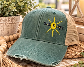 May include: A distressed green baseball cap with a tan mesh back. The front features an embroidered yellow sun graphic with the text Salt & Light in dark blue lettering. The brim has a frayed, worn-in edge and decorative stitching.