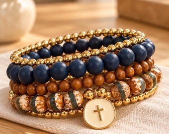 May include: A stack of four beaded bracelets featuring metallic gold spheres, matte navy blue beads, smooth brown wooden beads, and marbled brown and grey stone beads. A round cream-colored charm with a gold cross emblem hangs from the stack.