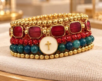 May include: A multi-strand beaded bracelet set featuring gold-toned metallic spheres, deep red square-cut glass beads, vibrant red faceted crystal beads, and dark teal round beads. A circular white charm with a gold cross is attached.