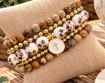 May include: A multi-strand beaded bracelet set featuring a mix of natural brown and tan stone beads, polished gold-tone metallic beads, and white ceramic beads decorated with small black floral patterns, centered by a round white charm with a gold cross.