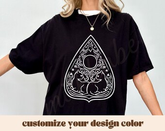May include: Black t-shirt featuring a white graphic of two cats within a decorative planchette design. The planchette includes stars, moons, and a central circle. The text "customize your design color" is displayed below the graphic.