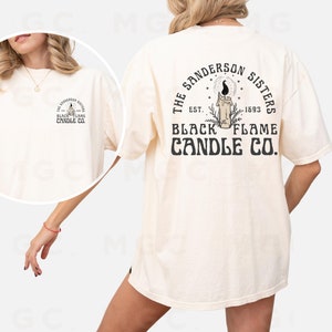 Pu&ograve; includere: Una t-shirt color crema con una stampa grafica sul retro che dice "The Sanderson Sisters Est. 1693 Black Flame Candle Co."