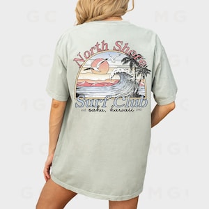 May include: A light green t-shirt with a graphic print of a sunset over a wave with palm trees and the text "North Shore Surf Club est. Oahu, Hawaii 1997".