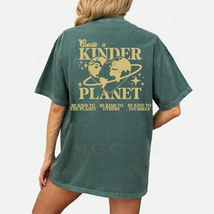 Peut inclure: Un t-shirt vert avec un graphique d'une plan&egrave;te en forme de c&oelig;ur avec le texte "Create a Kinder Planet" et "Be kind to the planet, be kind to others, be kind to yourself".