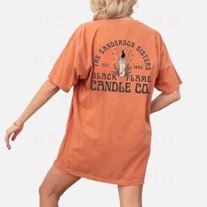Pu&ograve; includere: Una maglietta color arancio bruciato con una stampa grafica sul retro. La grafica presenta una candela nera con una fiamma e il testo "The Sanderson Sisters Est. 1693 Black Flame Candle Co."